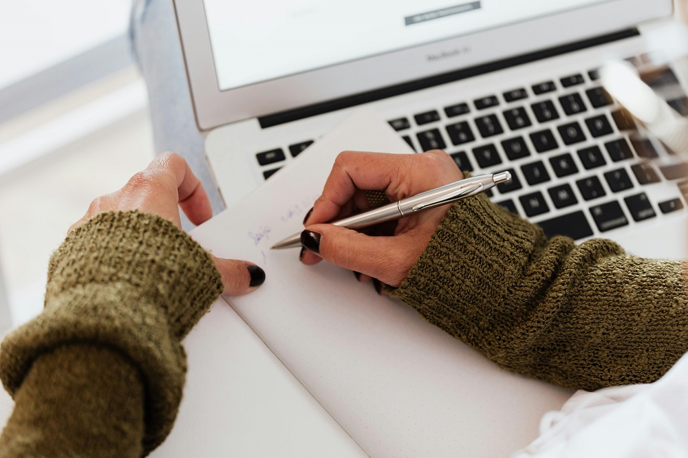 Person writing in a book in front of a laptop.